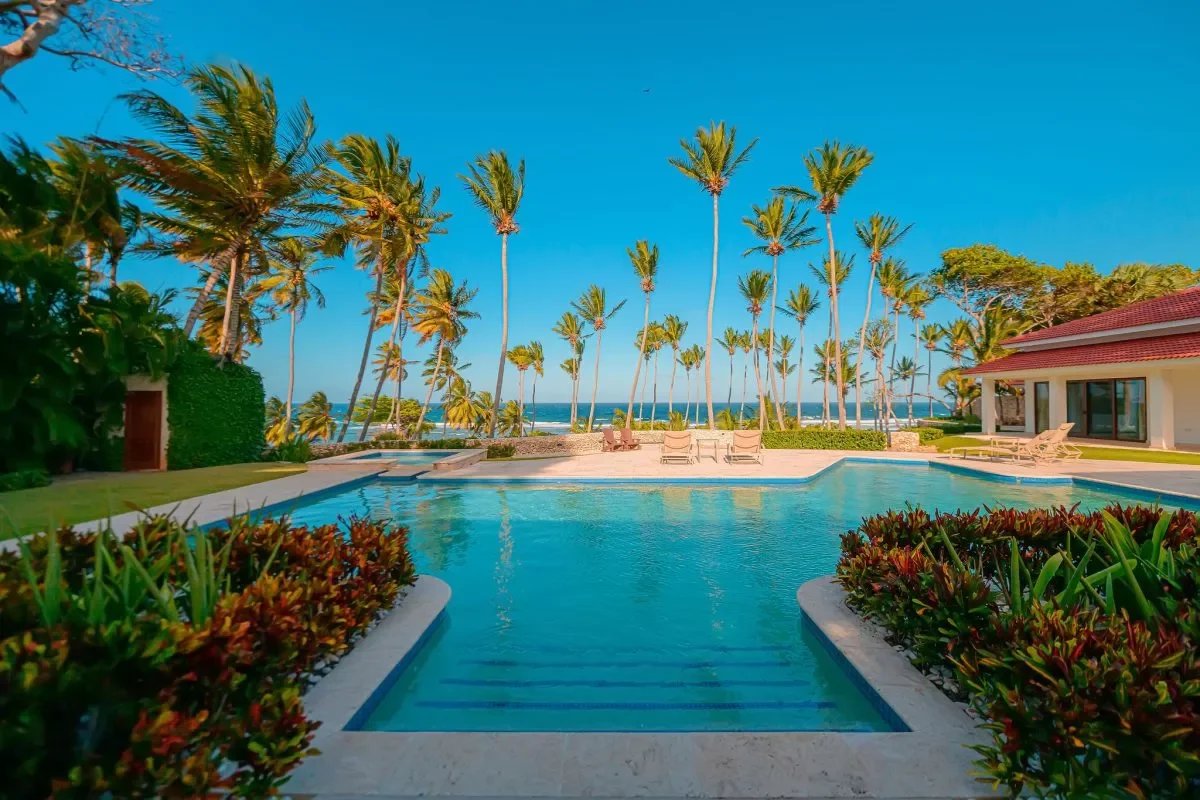 Resort pool with turquoise water, palm trees, and a view of the ocean beyond a lawn and lounge chairs.