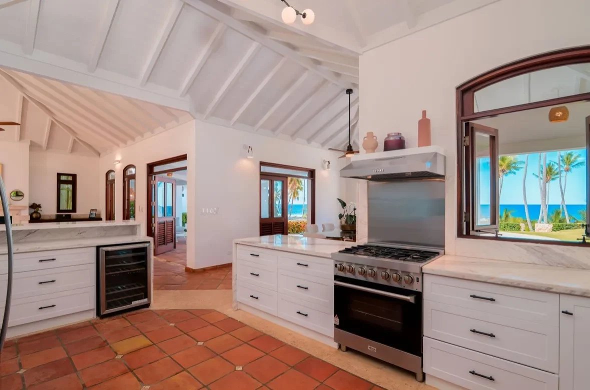 Bright coastal kitchen with white cabinets, marble counters, and a view of palm trees and the ocean through a large window.
