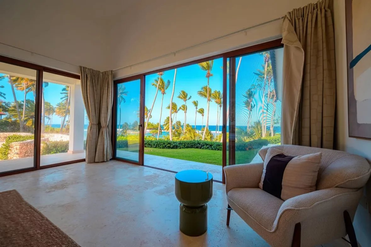 Living room with large glass sliding doors opening to a tropical garden and ocean view, beige sofa and cushions nearby.