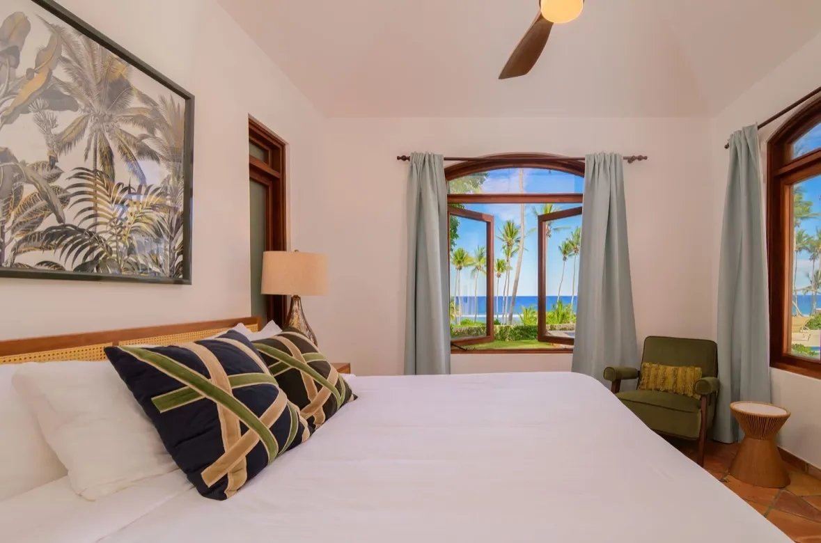 Bright hotel bedroom with a king bed, tropical pillows, and an open window to palms and the ocean.