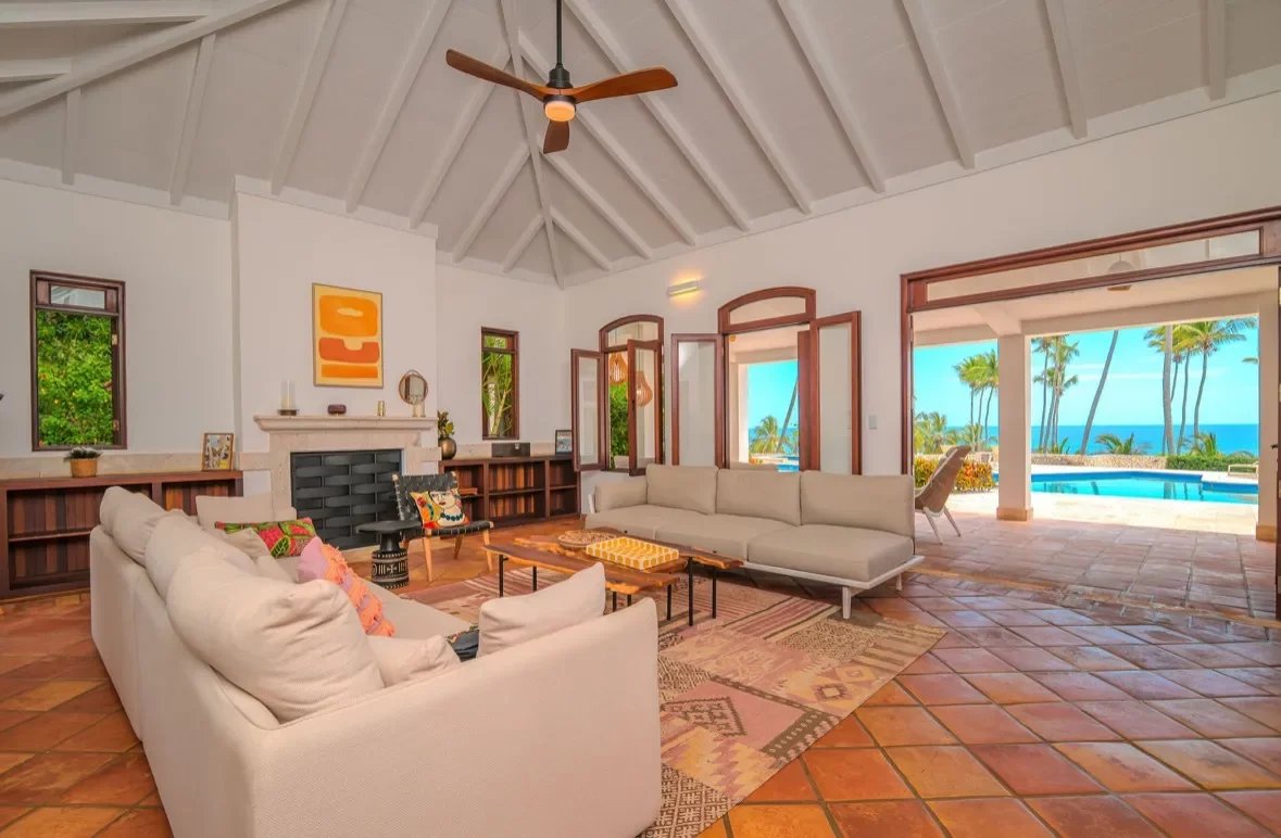 Spacious living room with white vaulted ceiling, beige sofas, and a view of the patio, pool, and ocean through open doors.