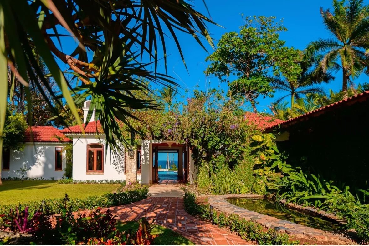 Tropical resort garden with a brick path, white building with a red roof, and an archway opening to the blue sea beyond.