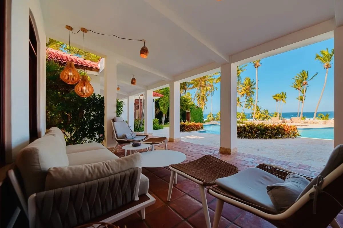 Covered patio with white cushioned seating and wicker loungers, overlooking a pool and palm-fringed beach.