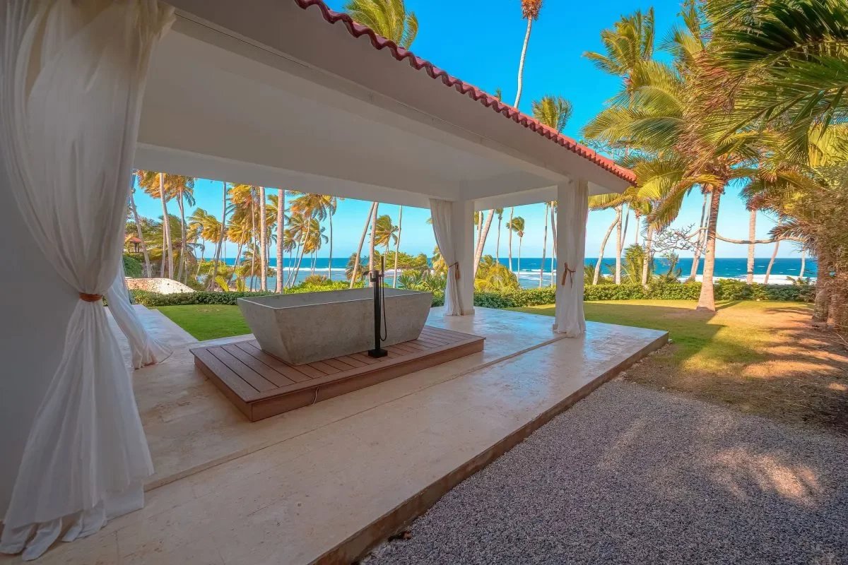 Open-air pavilion with white curtains and a stone bathtub on a wooden platform facing a tropical beach.