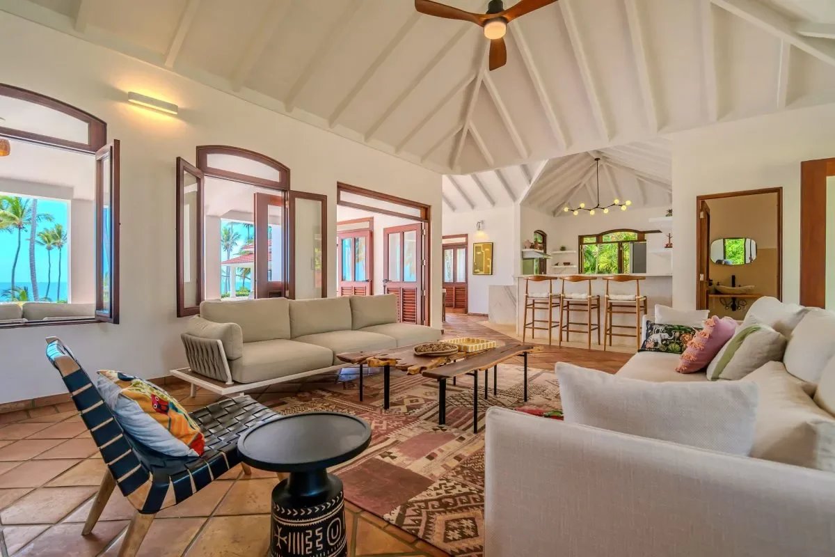 Bright tropical living room with white vaulted ceiling, beige sofa seating, and a bar area overlooking a beach view through open doors and windows.