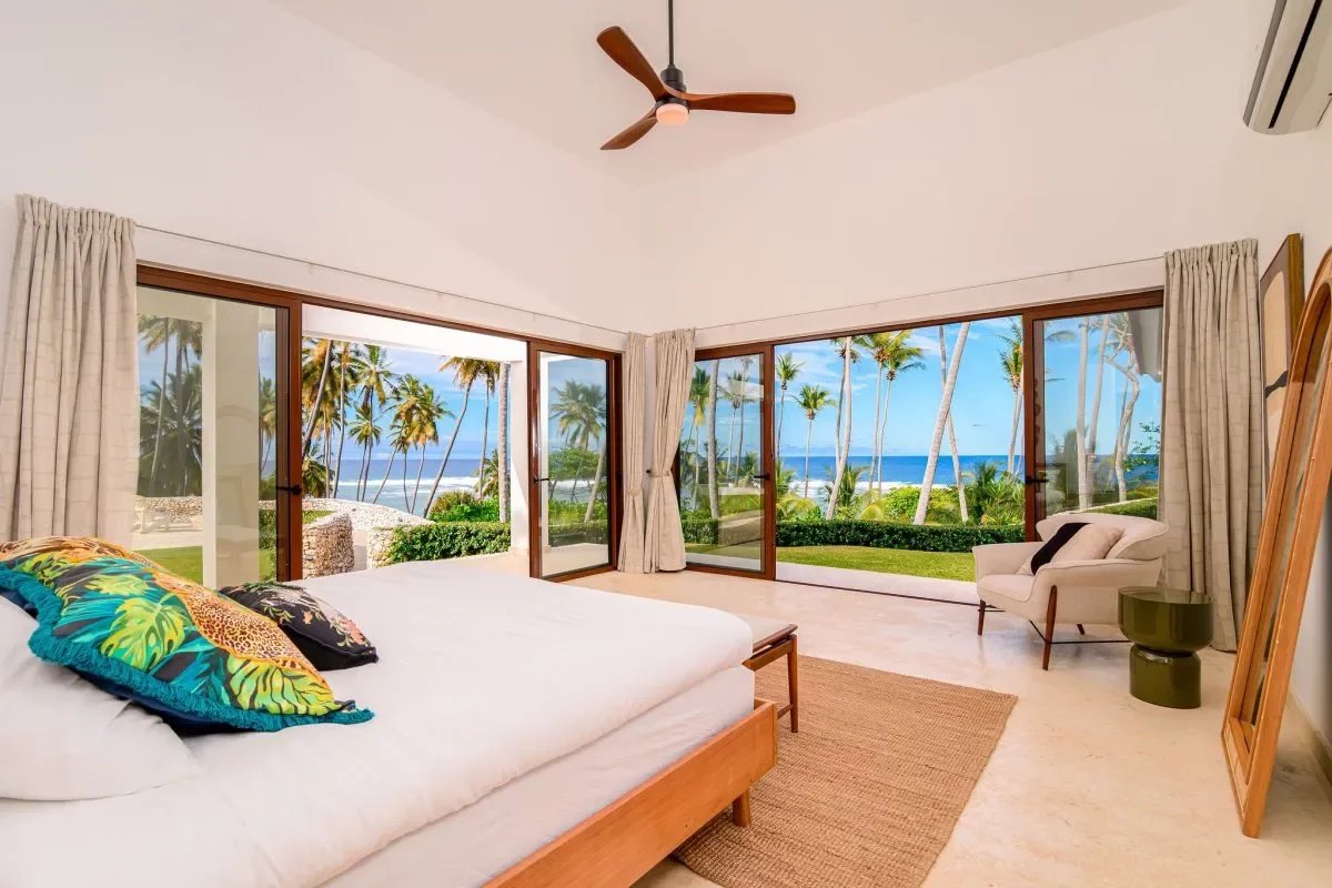 Sunlit tropical bedroom with a large bed, colorful pillows, and floor-to-ceiling glass doors opening to a palm-tree ocean view.