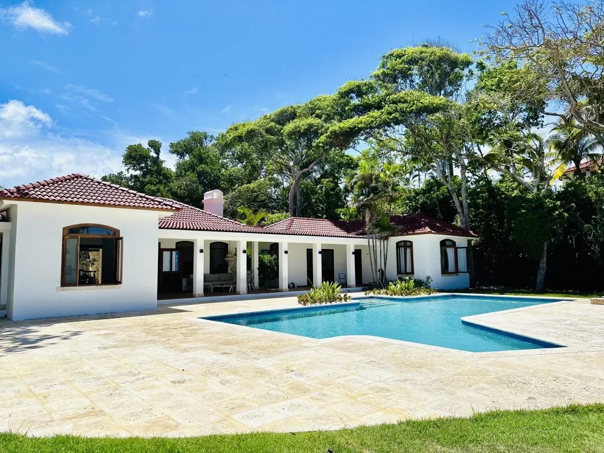 White single-story villa with a red tile roof, a covered patio, and a rectangular blue pool in a sunny backyard shaded by tall trees.