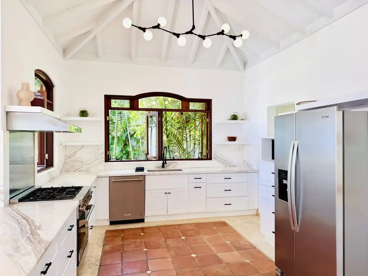 Bright white kitchen with marble countertops, stainless steel fridge and dishwasher, and a large window showing greenery outside.