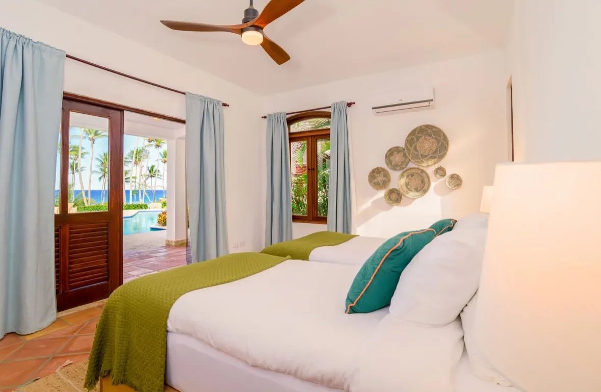 Bright hotel room with two white beds, green throws, and turquoise pillows; patio doors open to a seaside view with palm trees and water beyond.