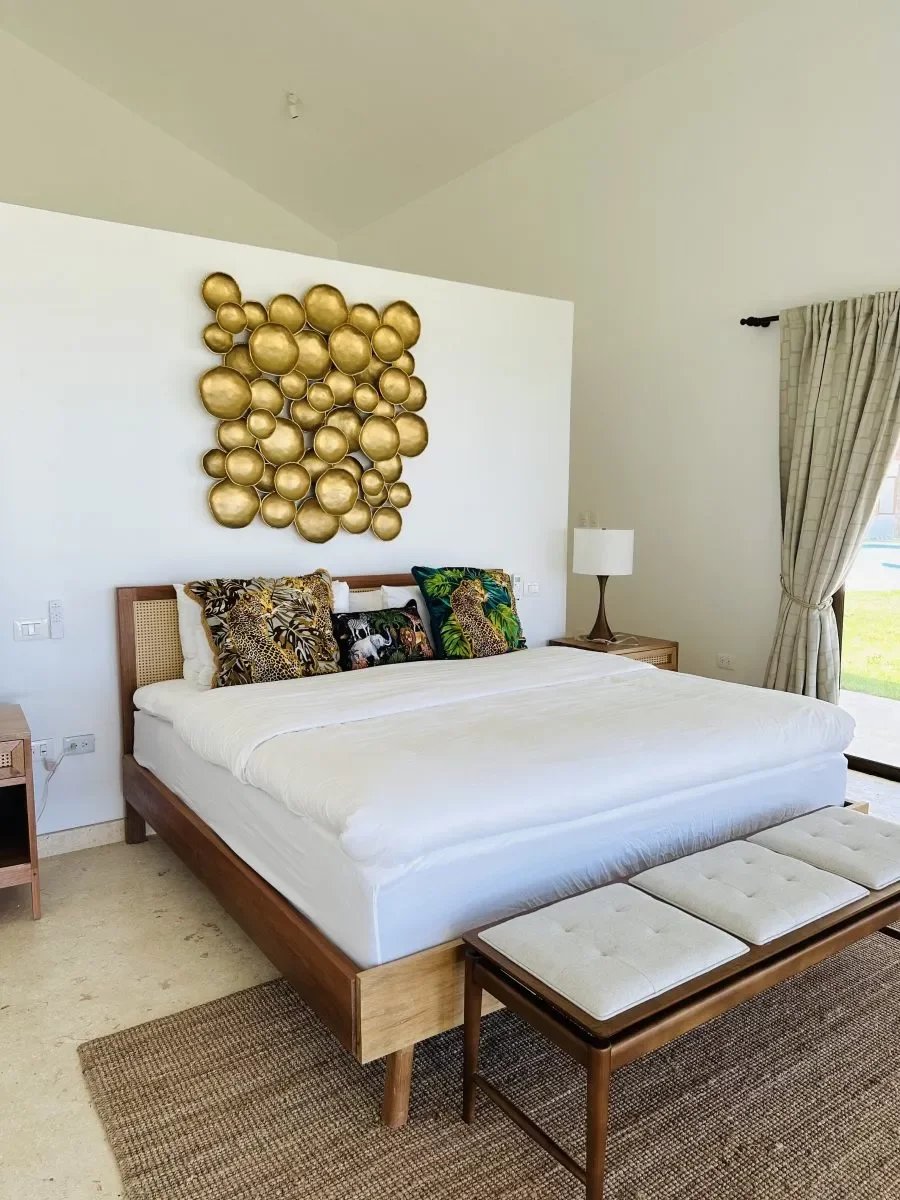 Hotel bedroom with a wooden platform bed, white bedding, gold circular wall art, tropical pillows, and a bench at the foot of the bed.