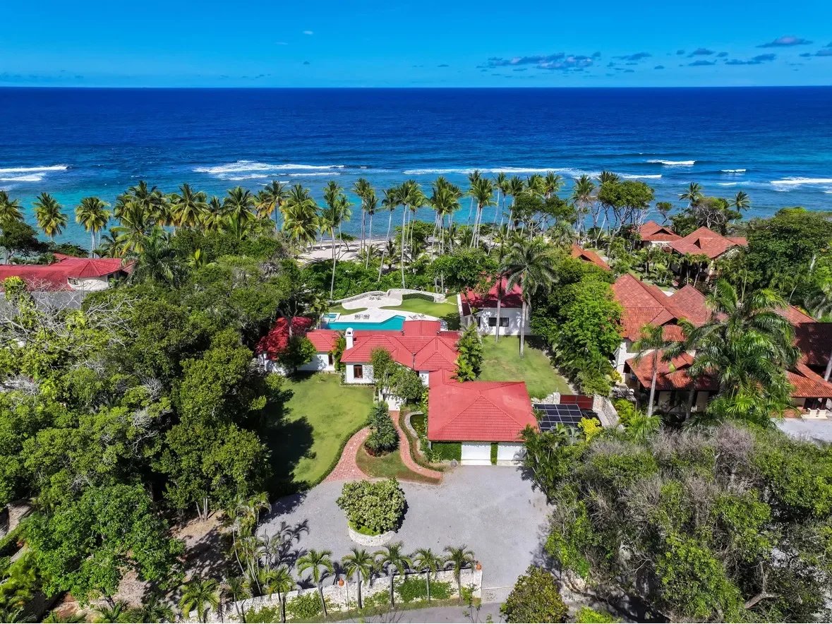 Aerial view of a tropical resort with red-roof villas, a central pool, and palm trees by the blue ocean coast.