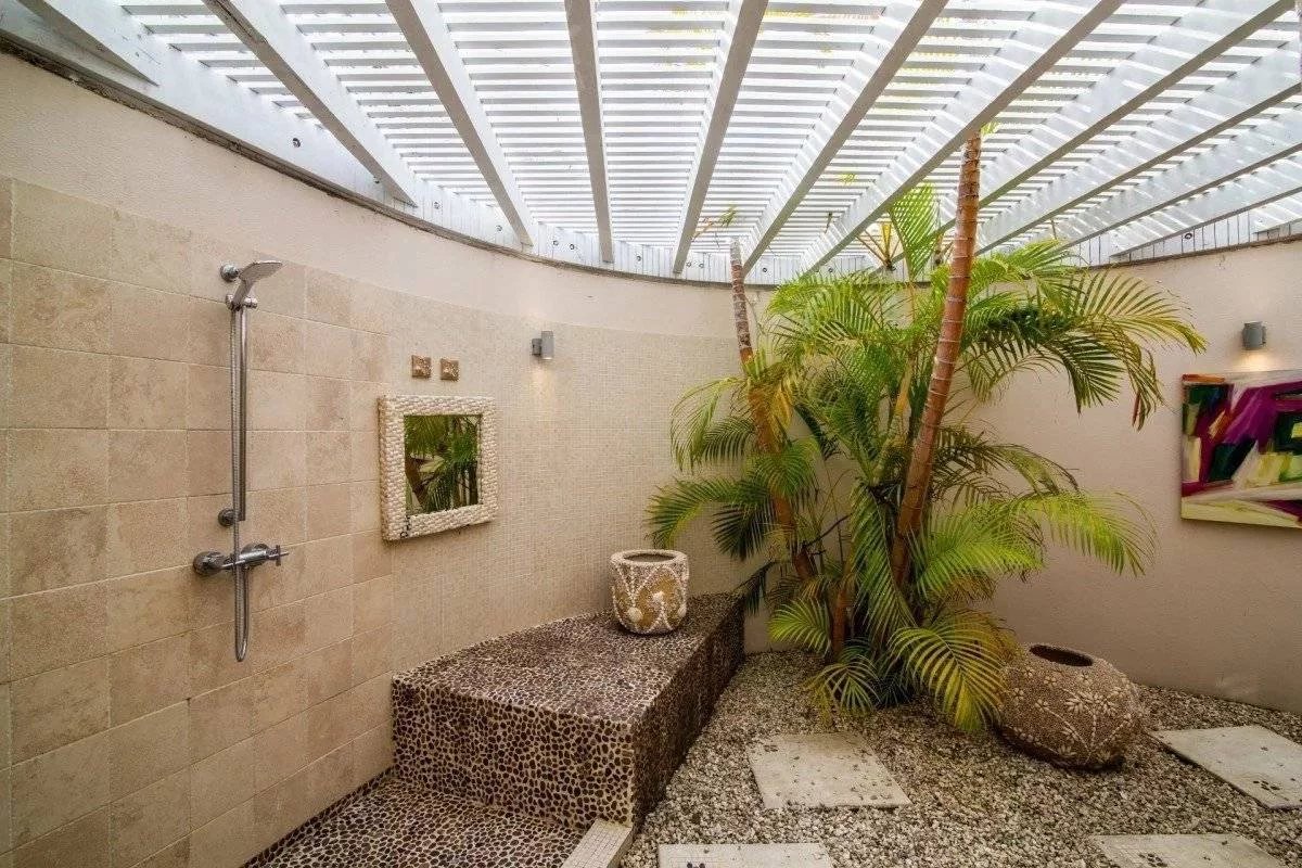 Indoor courtyard bathroom featuring a wall shower, beige tile walls, and pebble mosaic flooring with step platform. A lush palm plant fills the space under a translucent slatted roof.