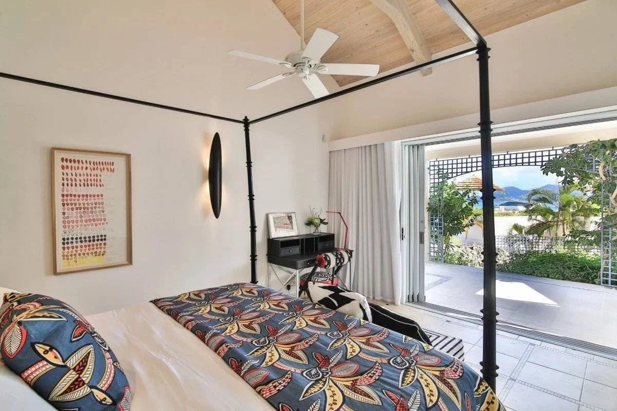 Bedroom with a black four‑poster canopy bed and tropical bedding, white walls, a ceiling fan, and a sliding door to a balcony with an ocean view.
