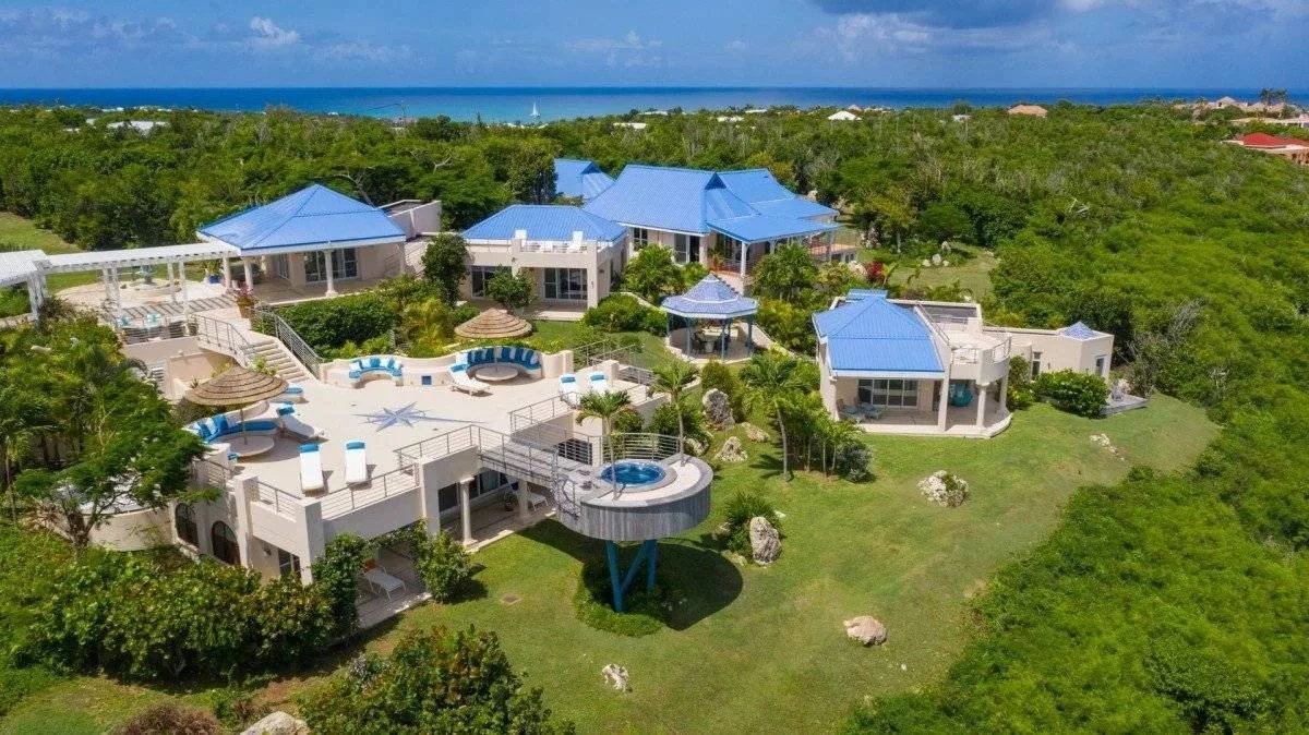 Aerial view of a white resort with blue roofs, multiple pools, and lush green surroundings near the ocean.