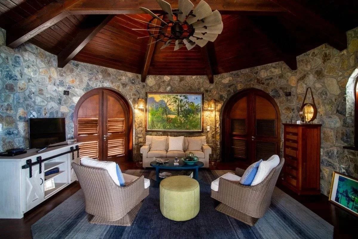 Rustic stone living room with arched wooden doors, beige sofa, and wicker chairs under a vaulted wood ceiling