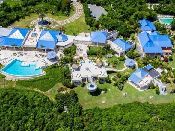 Aerial view of a luxury villa complex with blue-roofed buildings, multiple pools, and lush green grounds surrounded by trees.