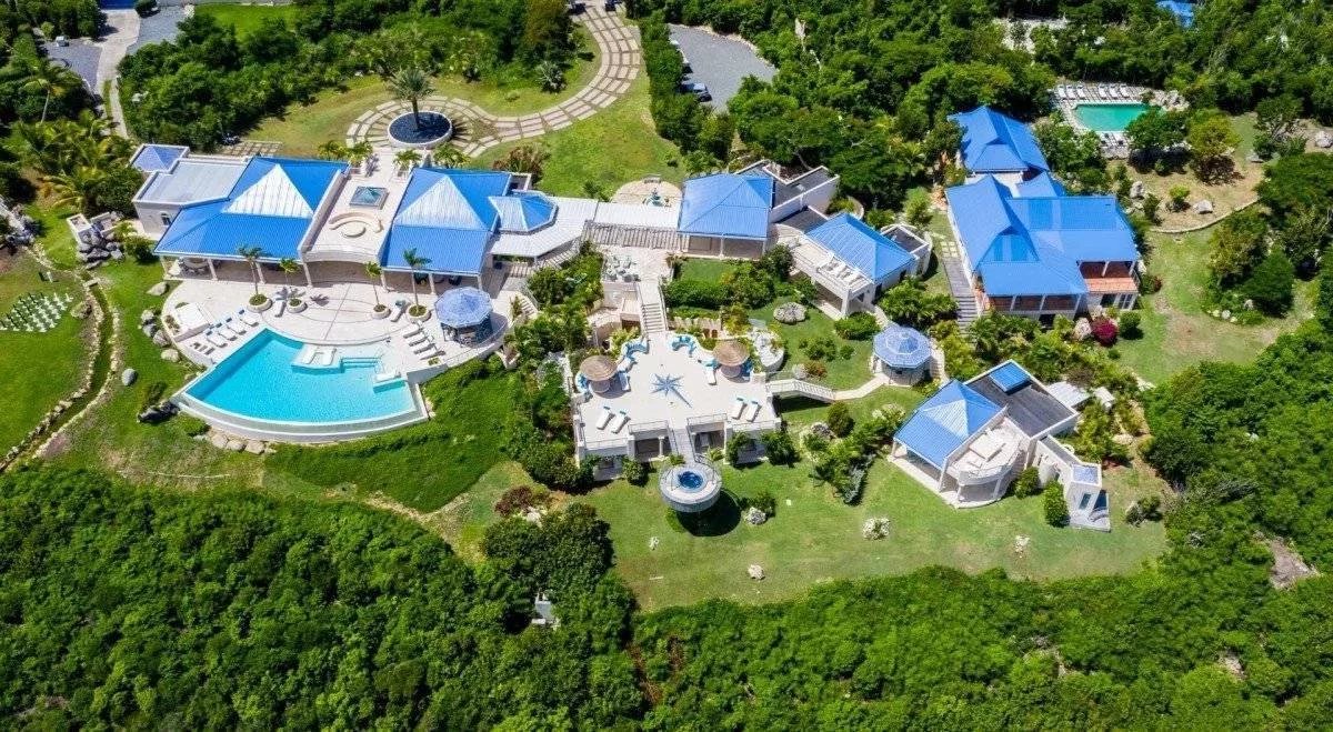 Aerial view of a luxury villa complex with blue-roofed buildings, multiple pools, and lush green grounds surrounded by trees.