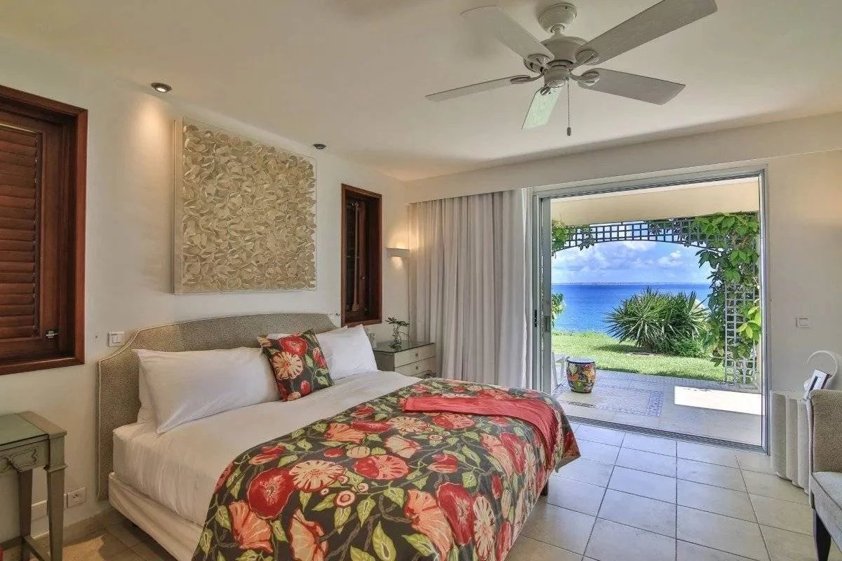 Bright bedroom with a tropical floral bedspread, white bedding, and a sliding door to a sea view outside.
