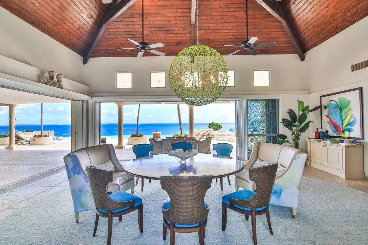 Open-plan dining area with a round glass table and blue-cushioned chairs, ocean view beyond sliding doors and a green spherical chandelier above.
