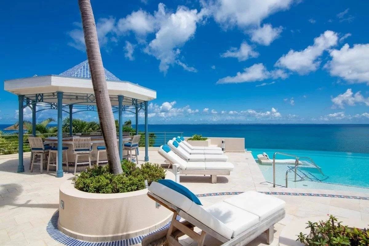 Poolside terrace at a tropical resort with white lounge chairs, a shaded bar area, and a palm tree, overlooking the blue ocean under a sunny sky.
