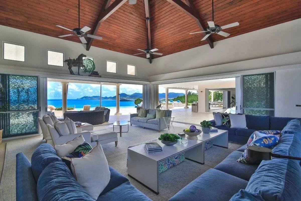 Open-concept living room with blue sectionals and white chairs, opening to an ocean-view patio through large sliding doors.