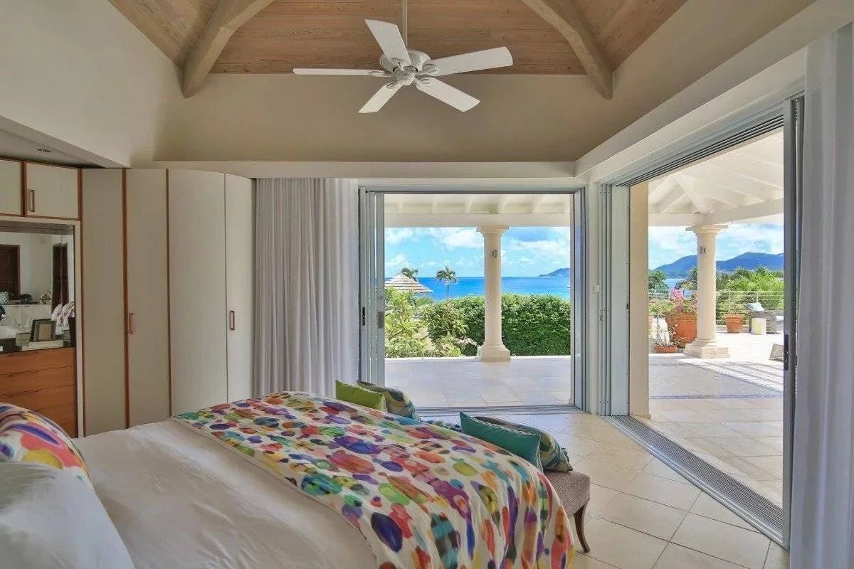 Bright beachfront bedroom with sliding glass doors opening to a terrace and ocean view; colorful quilt on the bed.
