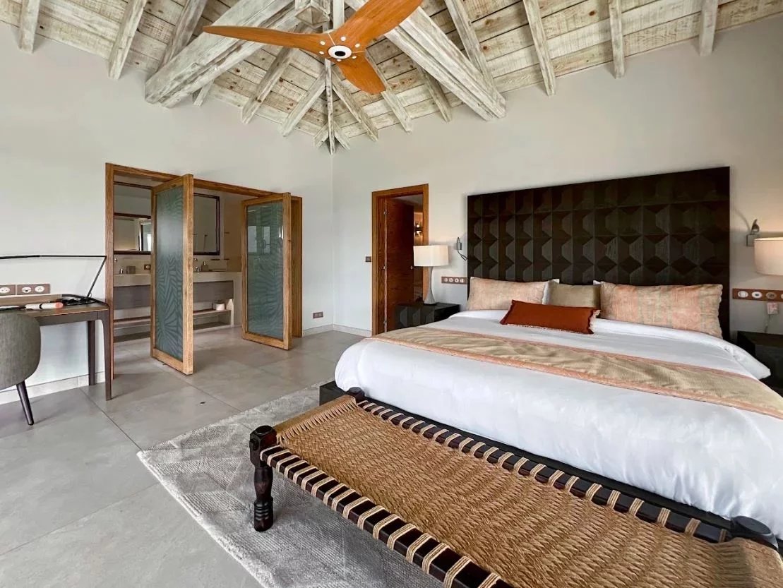 Luxurious hotel bedroom with a king bed, woven bench at foot, and exposed wooden beam ceiling; open doorway to a bathroom area.