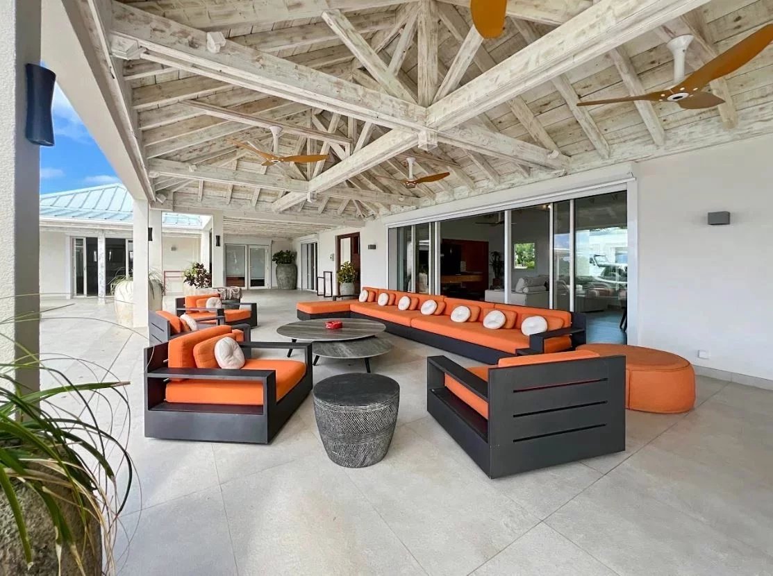 Outdoor covered patio with orange-cushioned modular seating and round coffee tables under a wooden beam ceiling.