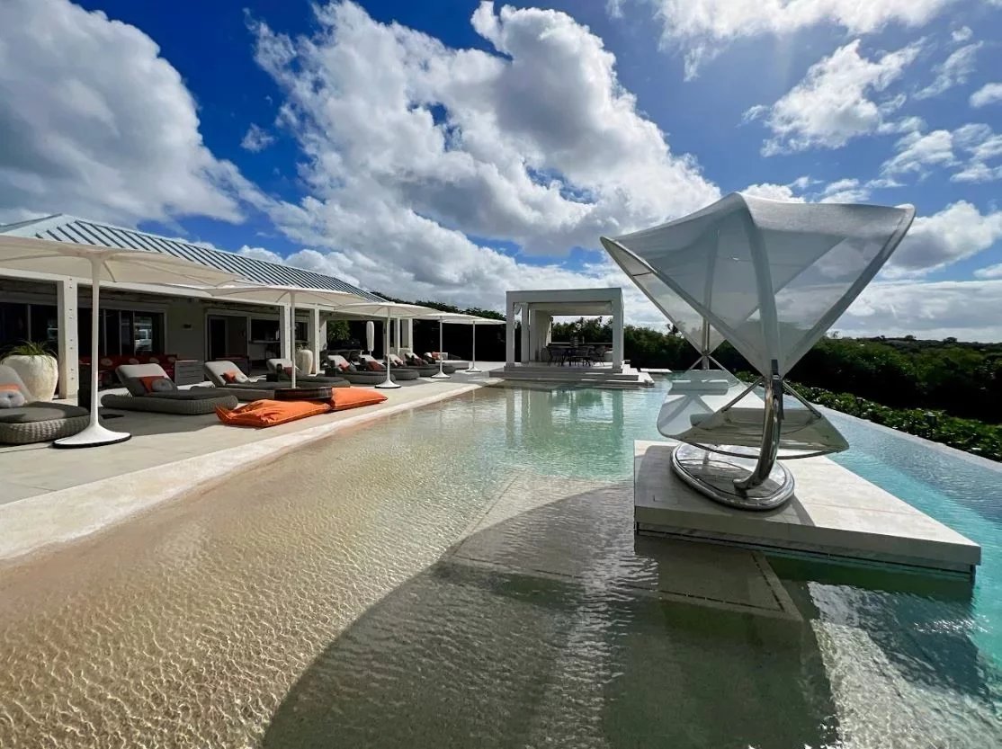 Luxurious poolside at a resort with white cabanas, lounge chairs, and a modern sculpture by the water under a blue sky.