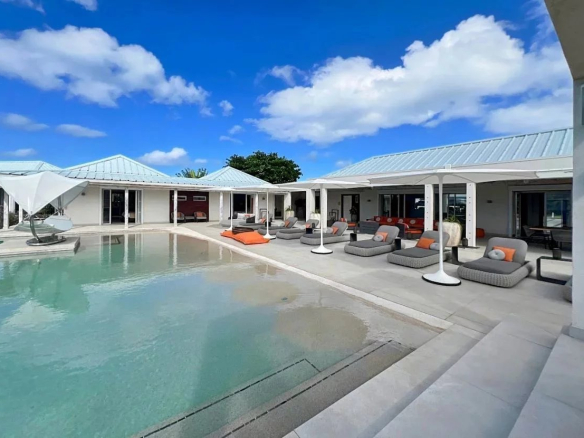 Poolside lounging area with gray chaise couches and orange pillows under white shade structures along a light-colored deck by a calm, turquoise pool.