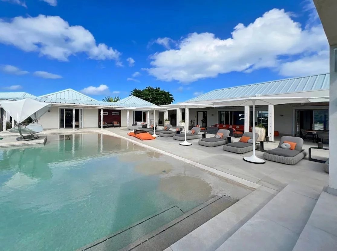 Poolside lounging area with gray chaise couches and orange pillows under white shade structures along a light-colored deck by a calm, turquoise pool.