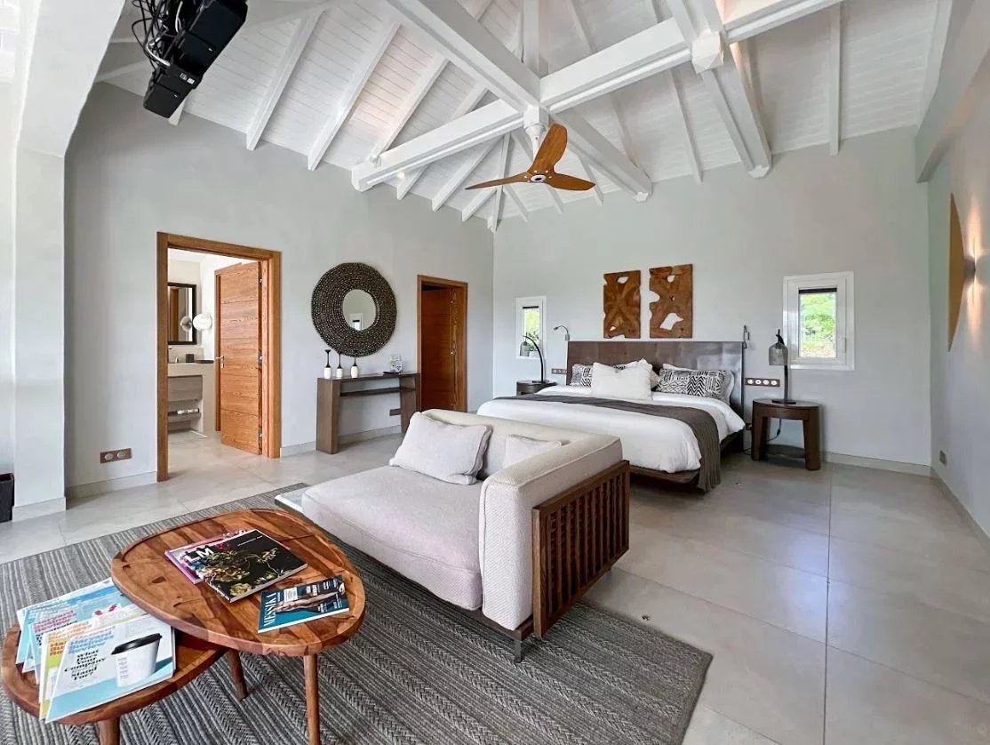Bright, airy bedroom with a vaulted white ceiling, king bed, and a seating area featuring two round wooden coffee tables and magazines on a rug.
