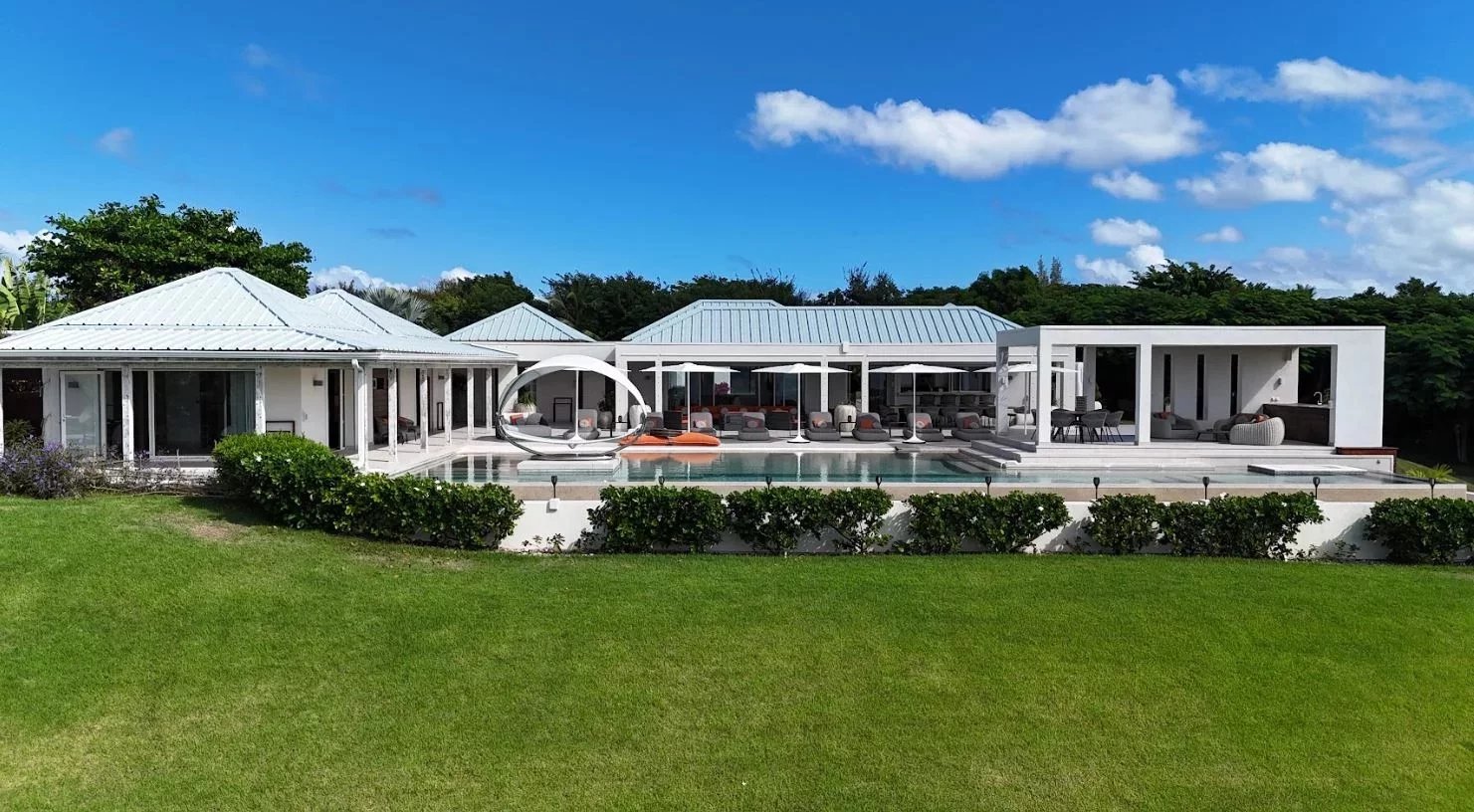 Luxurious white villa with multiple pavilions, a rectangular pool, and lounge areas on a sunny lawn.