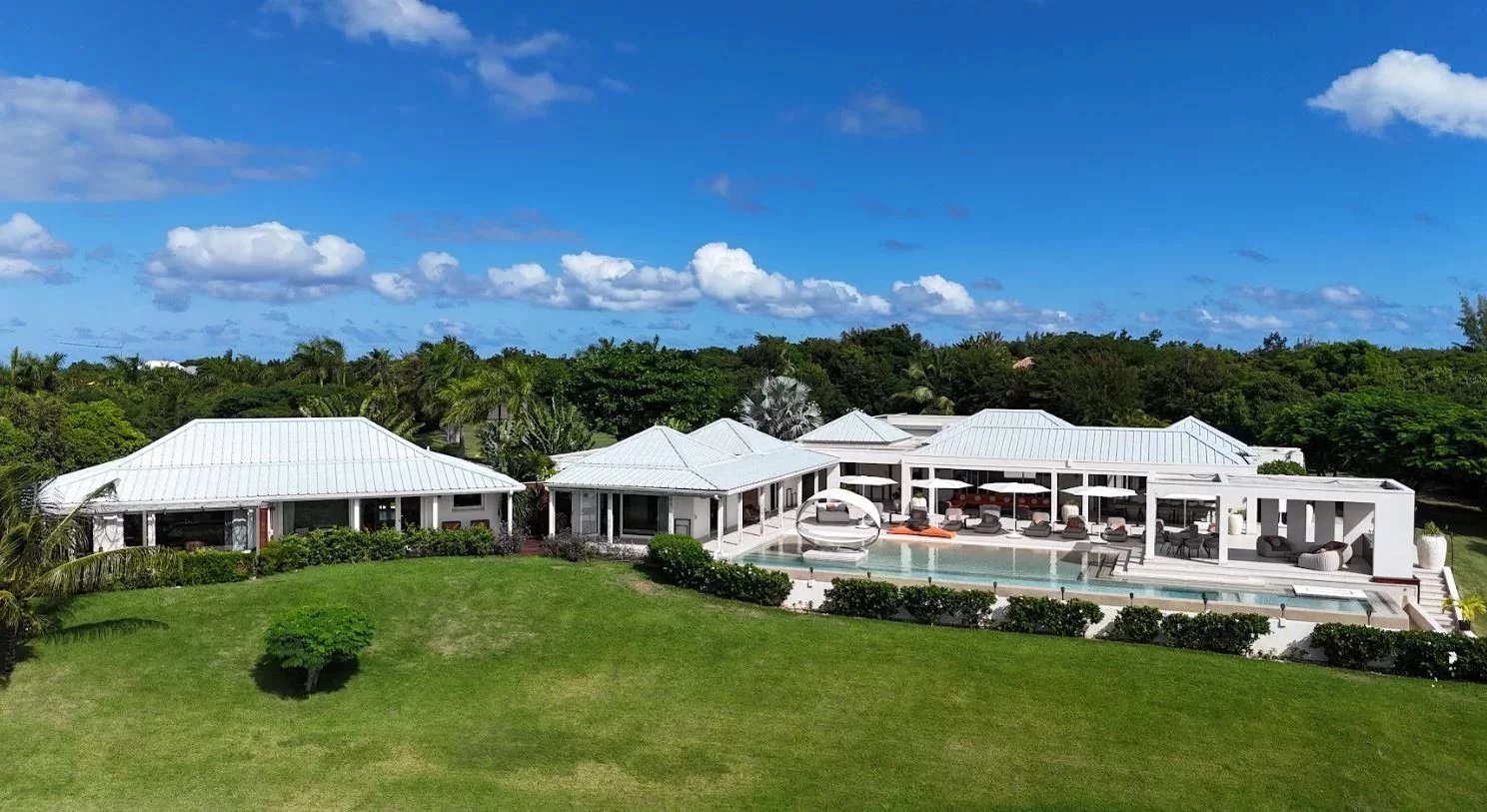 Aerial view of a white-roofed luxury villa with a long pool, loungers, and tropical landscaping under a blue sky.