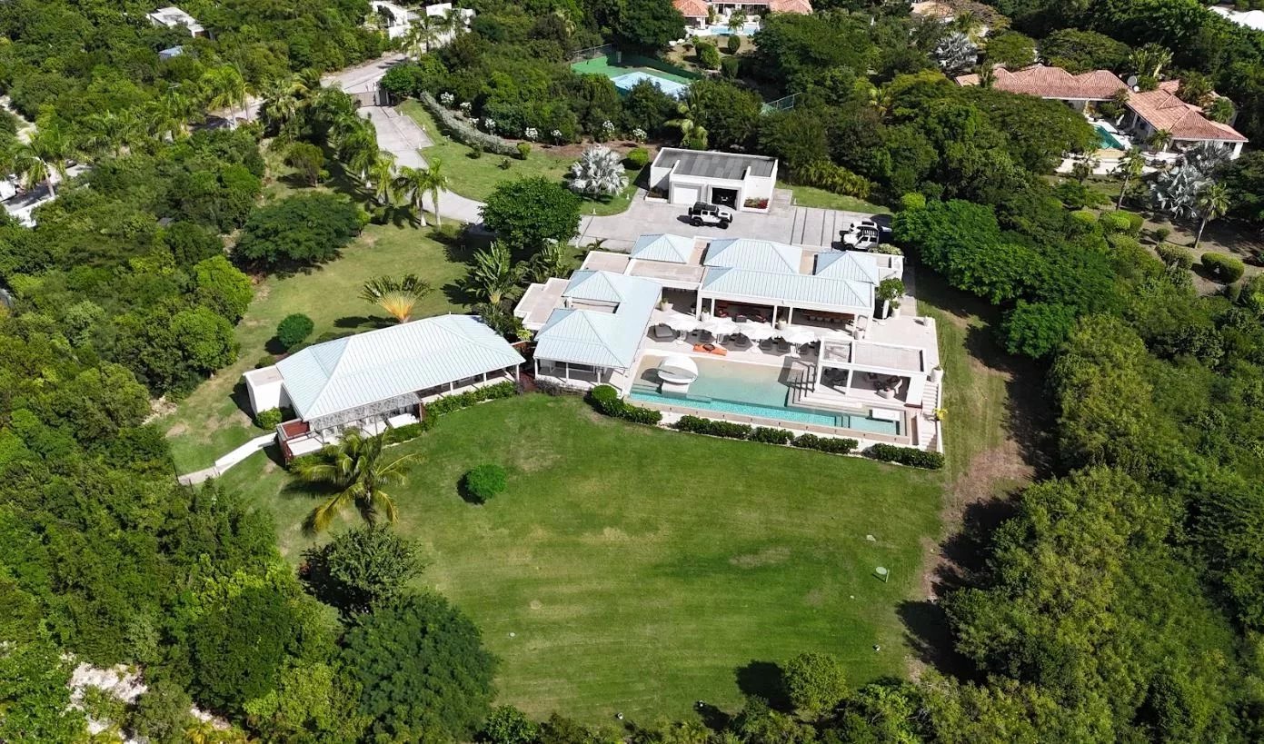 Aerial view of a large modern villa with white-roofed buildings, terraces, and a rectangular pool set in a lush green yard.