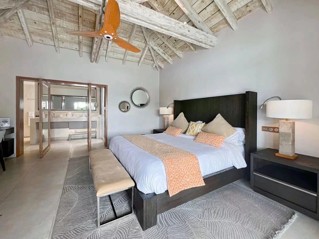 Hotel bedroom with a dark wooden king bed, orange runner and pillows, beige bench, and two nightstands with lamps.
