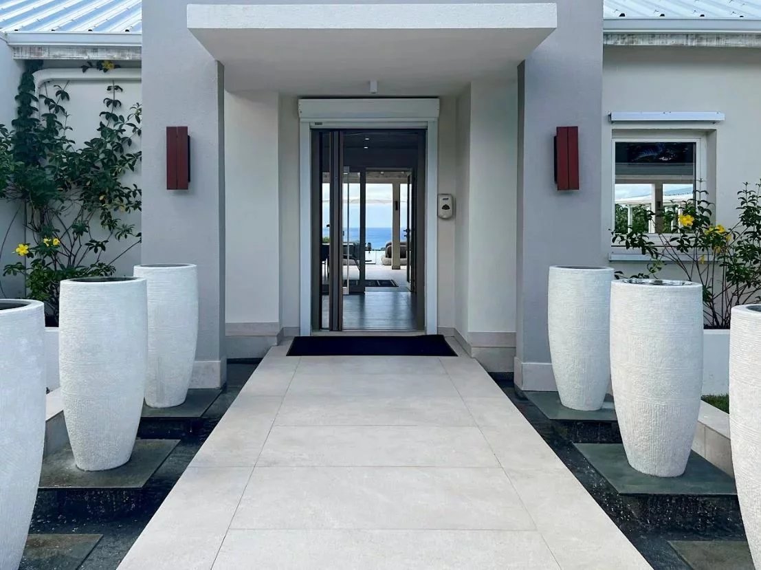 Modern entrance with a light tiled walkway, tall white planters, and glass doors opening to an ocean view beyond.