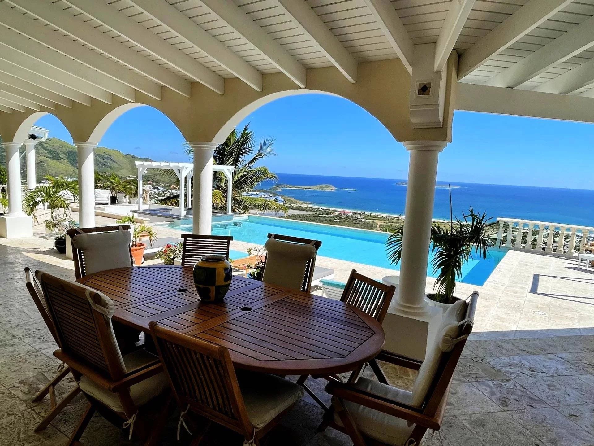 Covered patio with a round wooden dining table and cushioned chairs, arches and columns, overlooking a pool and the blue ocean.