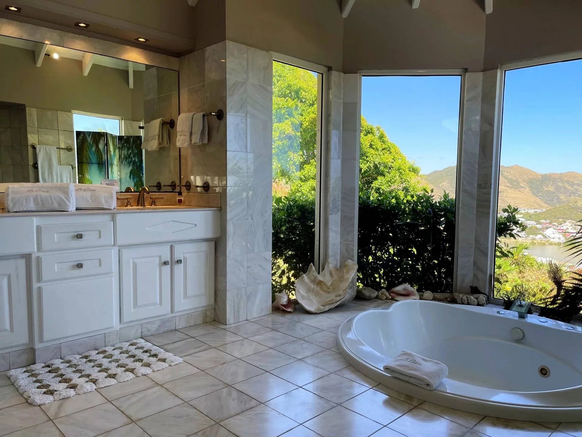 Bright bathroom with white cabinets, marble tile, and a large corner tub facing a garden and mountains outside.