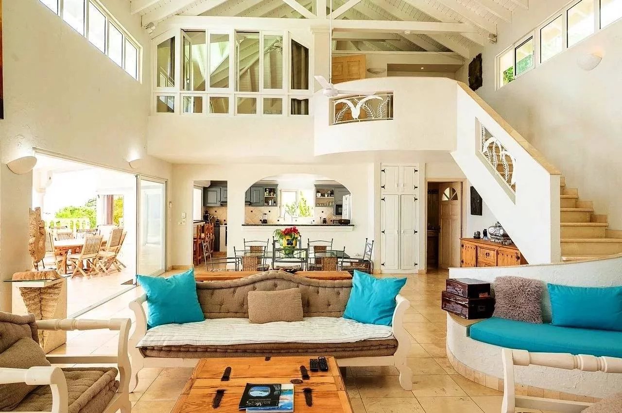 Bright open-plan living room with white furniture, turquoise cushions, and a view into the kitchen and dining area behind it.