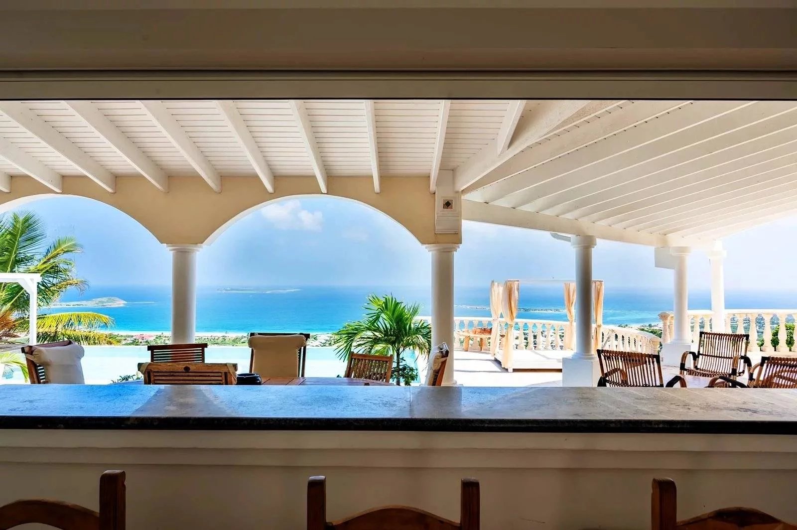 Open-air beachfront terrace with white arches and columns, overlooking the blue sea and palm trees, with wooden seating.",