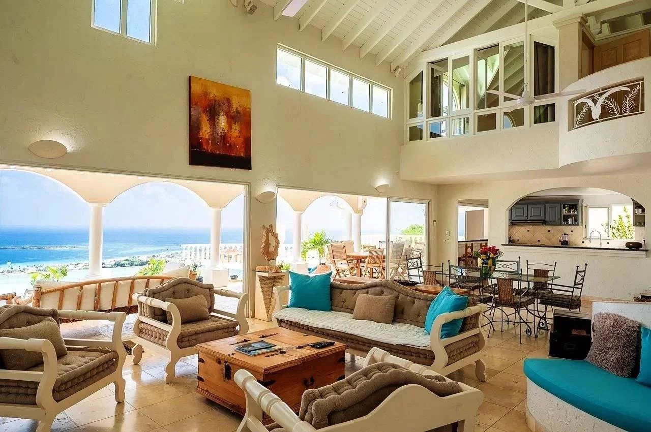 Bright beachfront living room with white furniture, turquoise cushions, and an ocean view through arched windows.