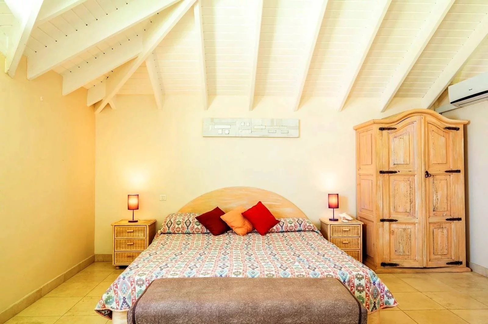 Cozy bedroom with a pitched ceiling, a wooden headboard, patterned bedspread, two nightstands with lamps, and a tall wooden armoire on the right side.