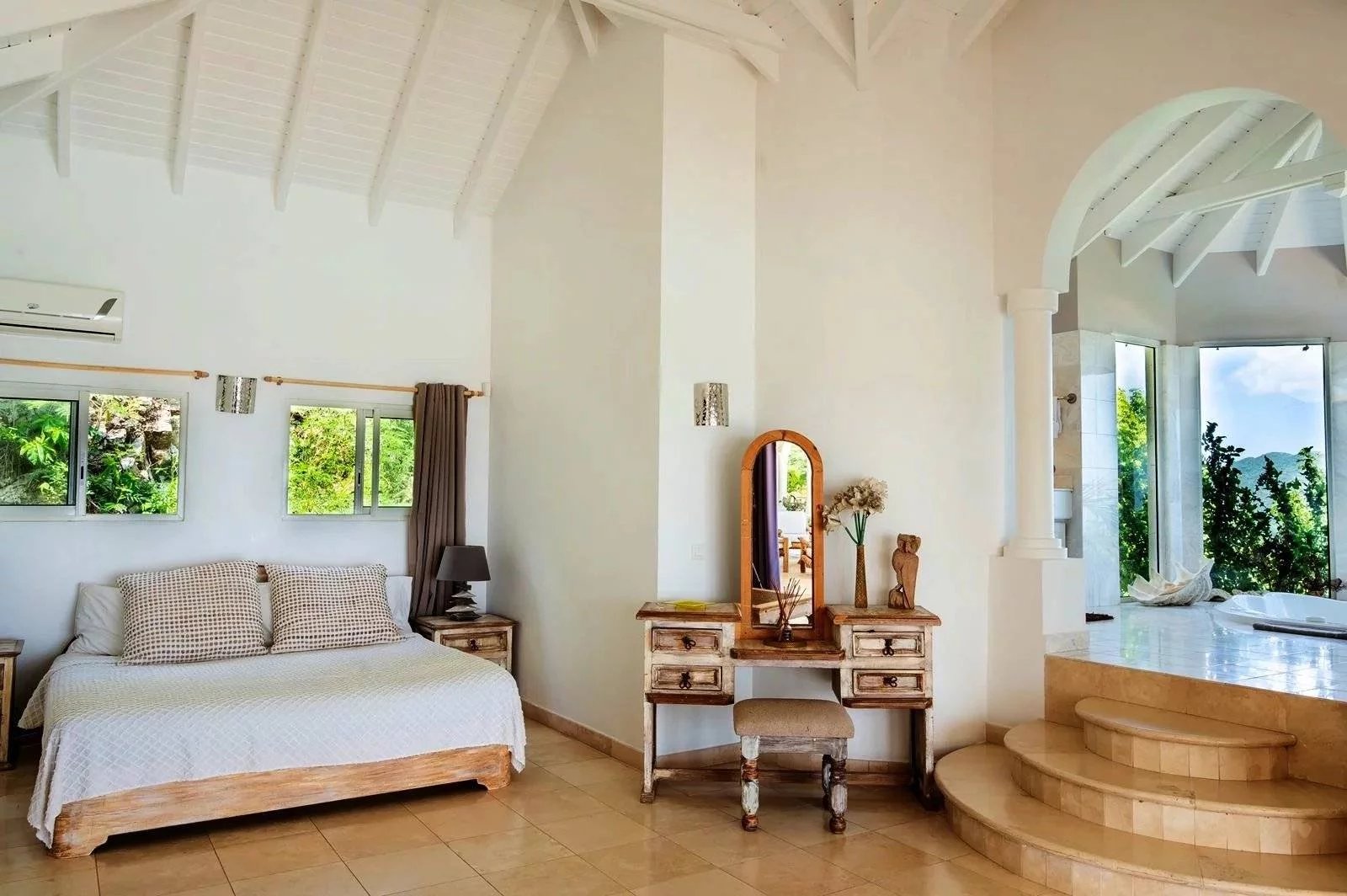 Bright bedroom in a white villa with a vaulted ceiling, wooden bed, and a vintage vanity desk with mirror.