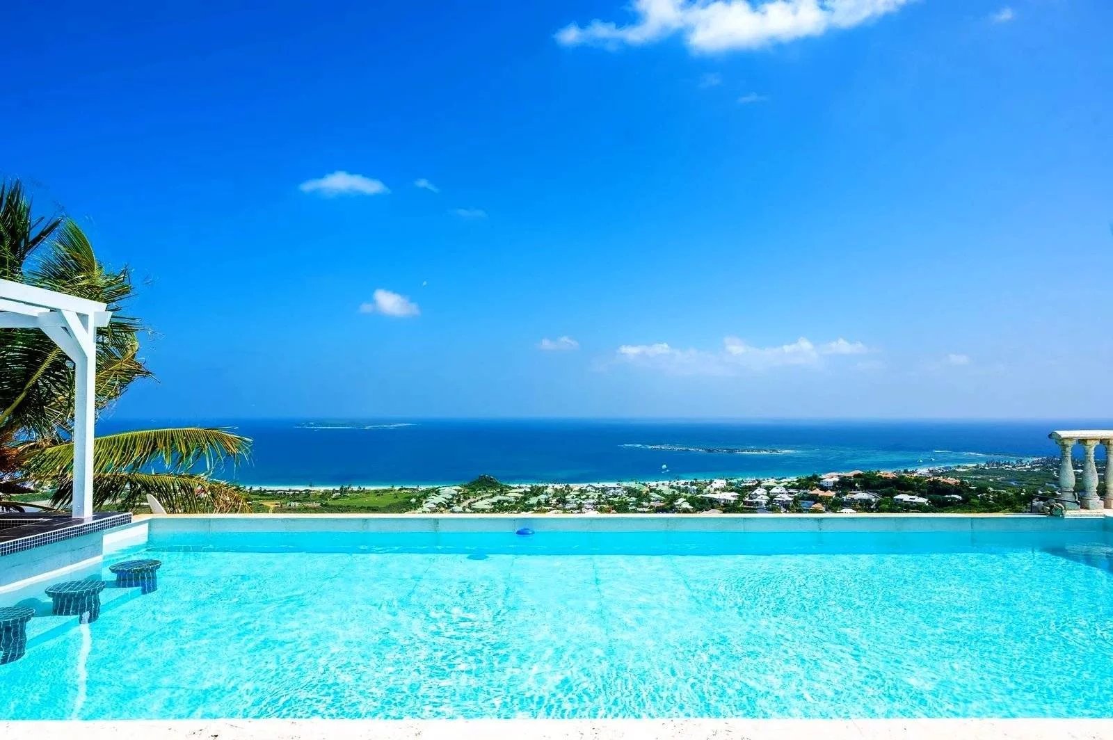 Infinity pool with clear blue water, palm trees, and a sea horizon under a bright blue sky.