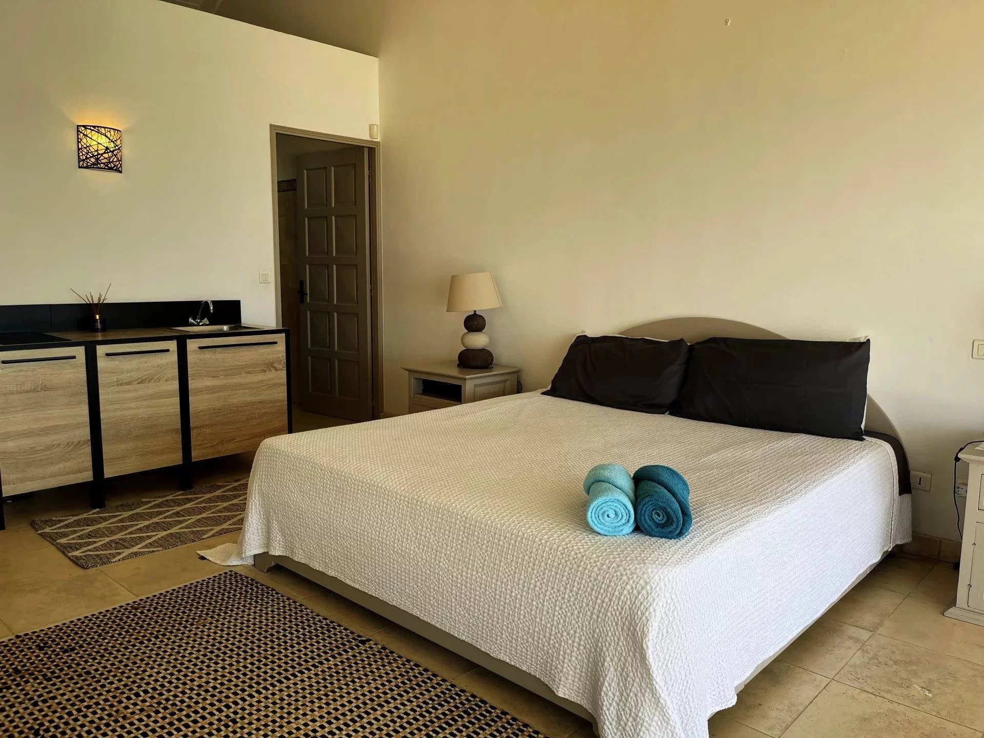 Hotel bedroom with a large white bed, two black pillows, and turquoise towels on top; bedside lamp and nightstand to the left, kitchenette along left wall, door in background.