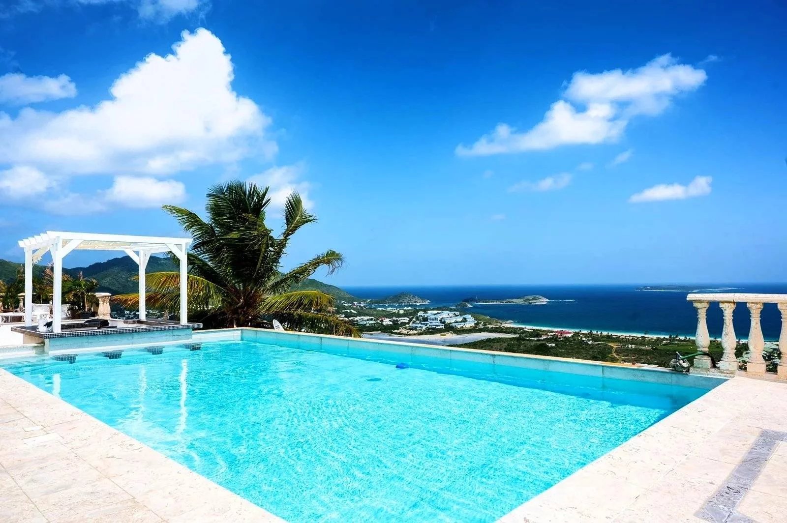 Infinity pool overlooking the blue sea with palm trees and a white pergola on a sunny day.