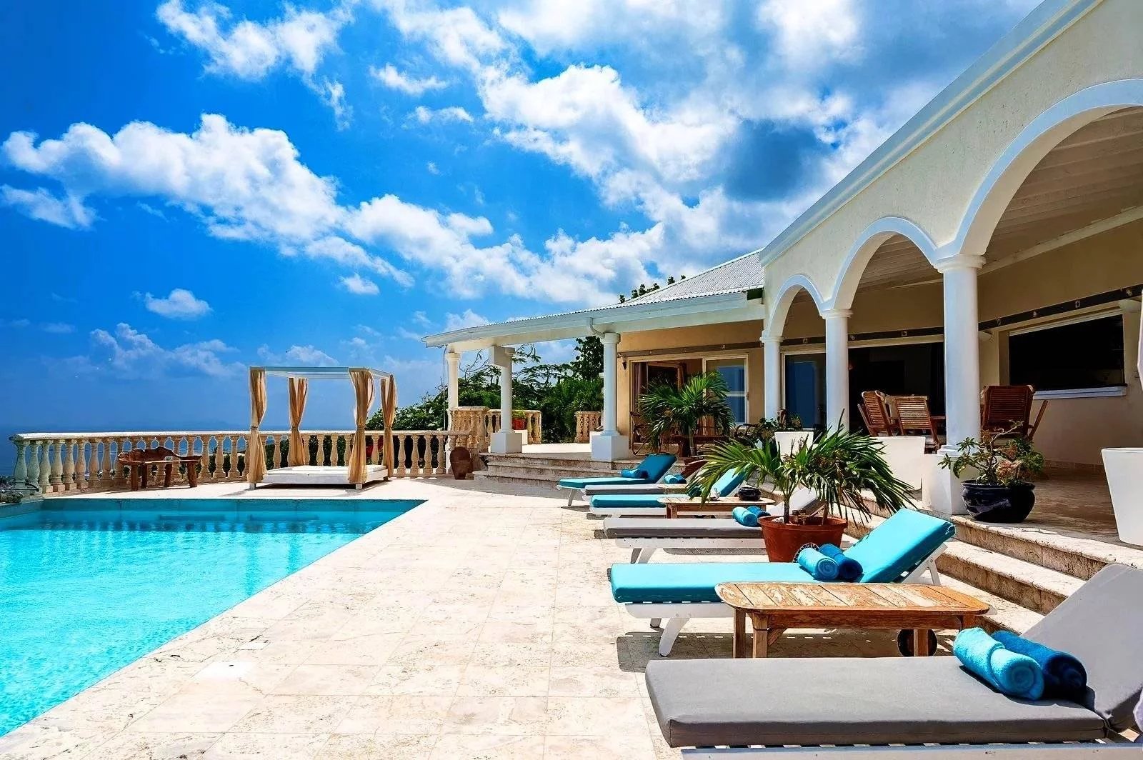 Sunny poolside terrace at a villa, with white arched columns, blue-cushioned loungers, and potted palms under a bright blue sky.
