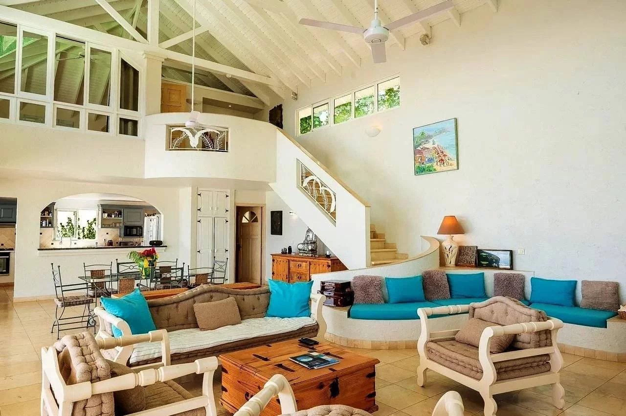 Bright open-plan living room with white furniture, turquoise cushions, and a curved staircase leading to a mezzanine; kitchen visible in the background.