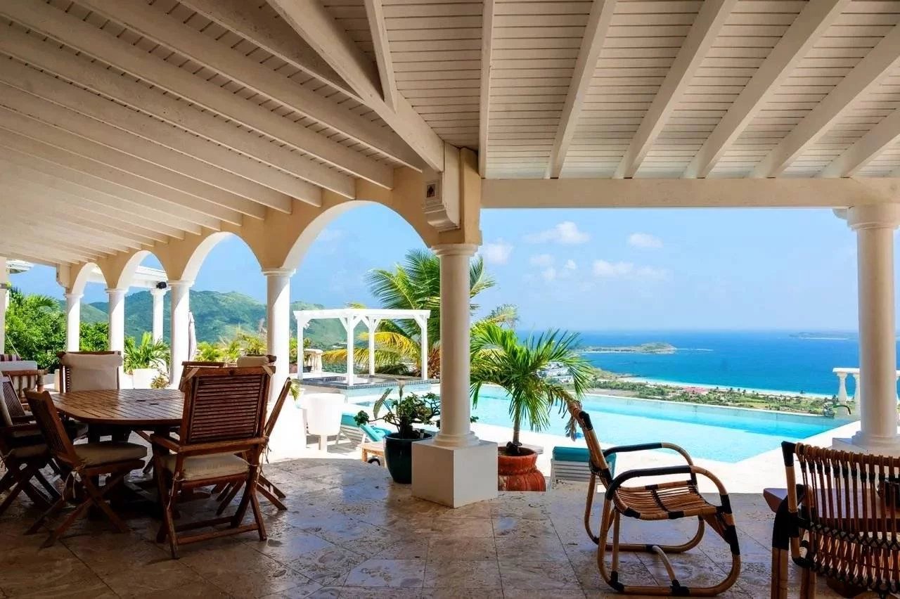 Open-air patio with wooden chairs around a table, overlooking a pool and turquoise sea beyond.